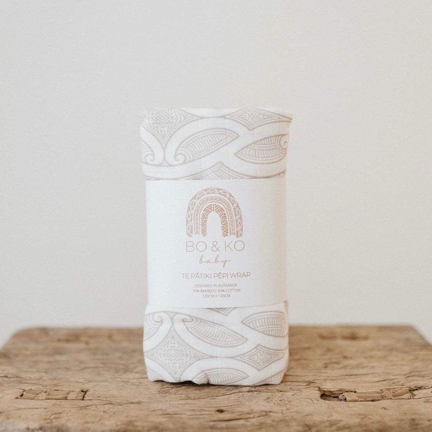 Bo & Ko Te Patiki Pepi Wrap in Mocha, rolled and banded on a wooden table