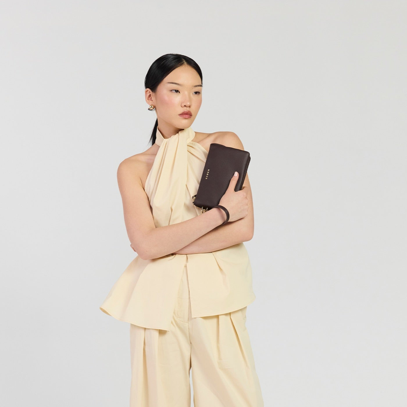 A model stands against a light gray studio background wearing a cream-colored asymmetrical halter top and matching wide-leg trousers, holding a dark brown clutch.