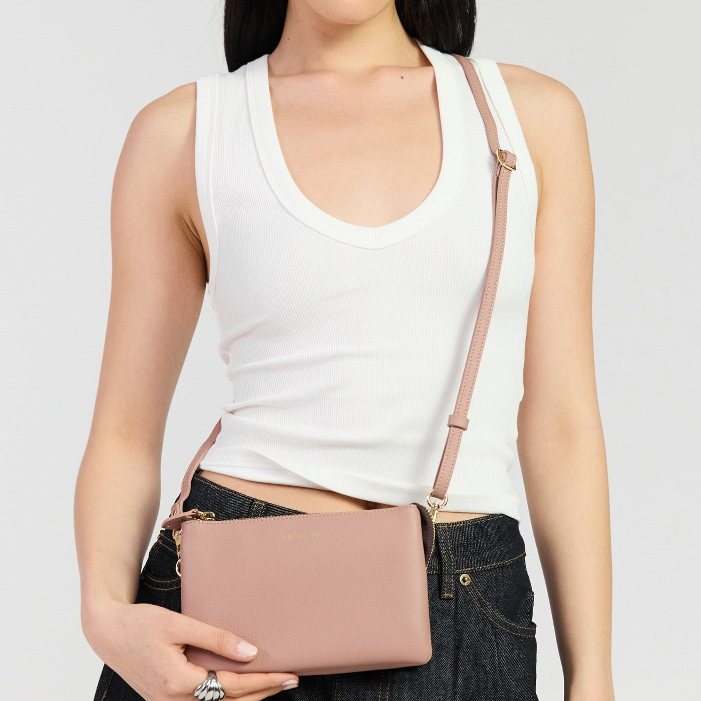 A model wearing a white ribbed tank top and dark denim jeans holds a dusty pink faux-leather crossbody bag with a matching shoulder strap against a light gray studio background.