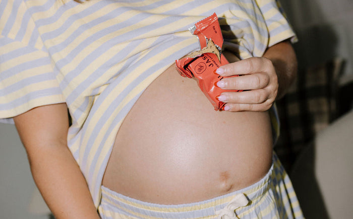 A pregnant woman wearing a light yellow and white striped t-shirt and matching drawstring shorts, lifting her shirt to show her baby bump while holding a small orange pouch.